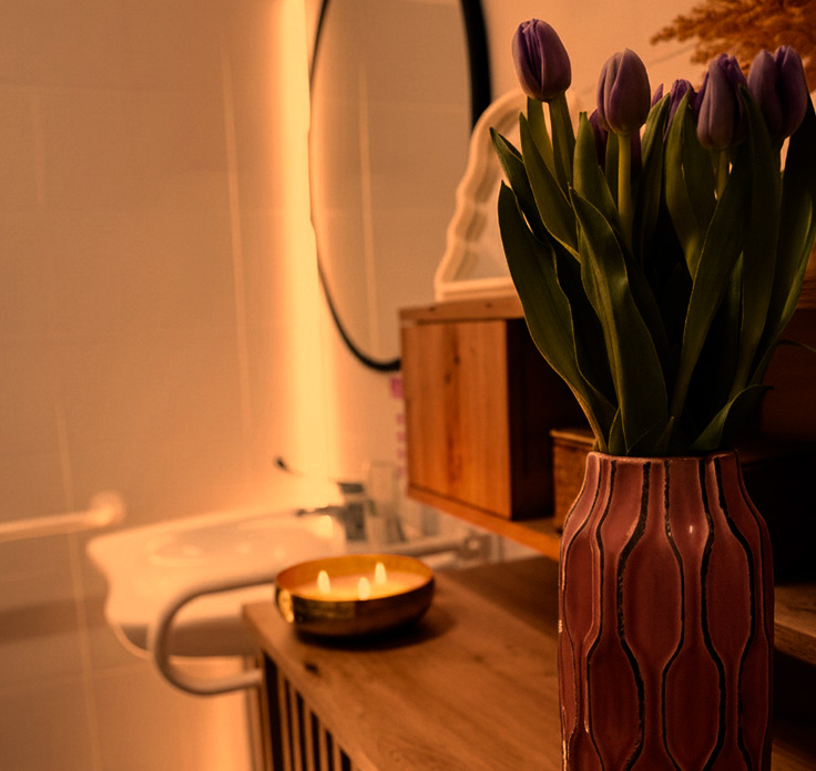 Wazon z fioletowymi tulipanami na stole; w tle umywalka, świeczka w drewnianej podstawce i owalne lustro.